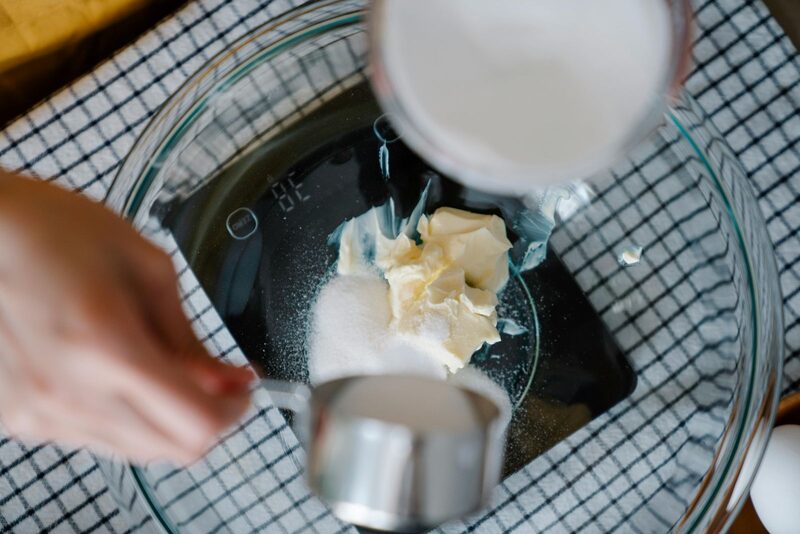 Hand pouring sugar onto a digital kitchen scale holding vegan butter and flour in a glass bowl