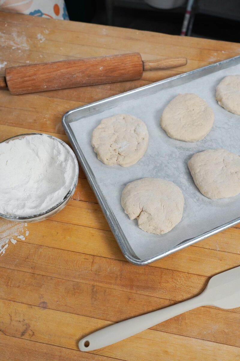 Cut dough rounds arranged on a parchment-lined baking sheet beside a bowl of flour and a rolling pin