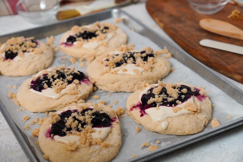 Unbaked buns topped with cheesecake swirl, blueberry filling, and a crumb topping before baking