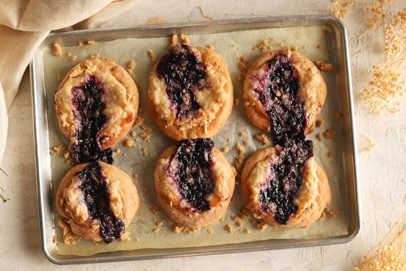A parchment-lined sheet pan of six baked blueberry cheesecake buns topped with cheesecake swirl and blueberry filling