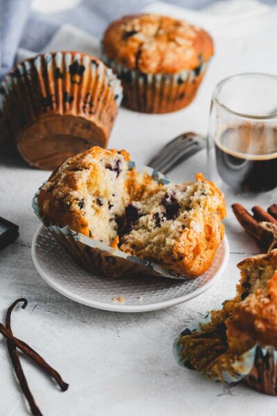 A blueberry crumb muffin cut in half on a plate with more muffins and coffee around