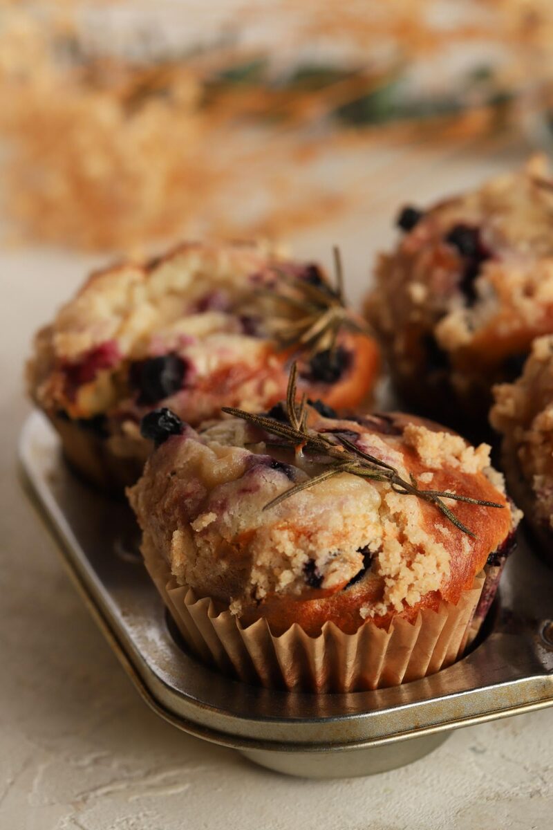A single blueberry rosemary muffin in a brown paper liner, close up, with a crumbly streusel top and a sprig of fresh rosemary