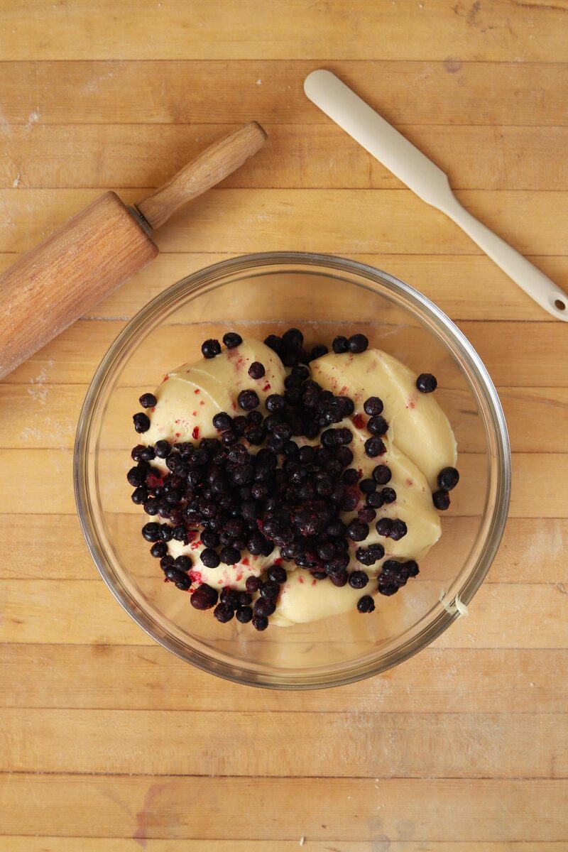 Blueberries piled on pale muffin batter in a glass bowl, ready to be folded in, beside a rolling pin and spatula