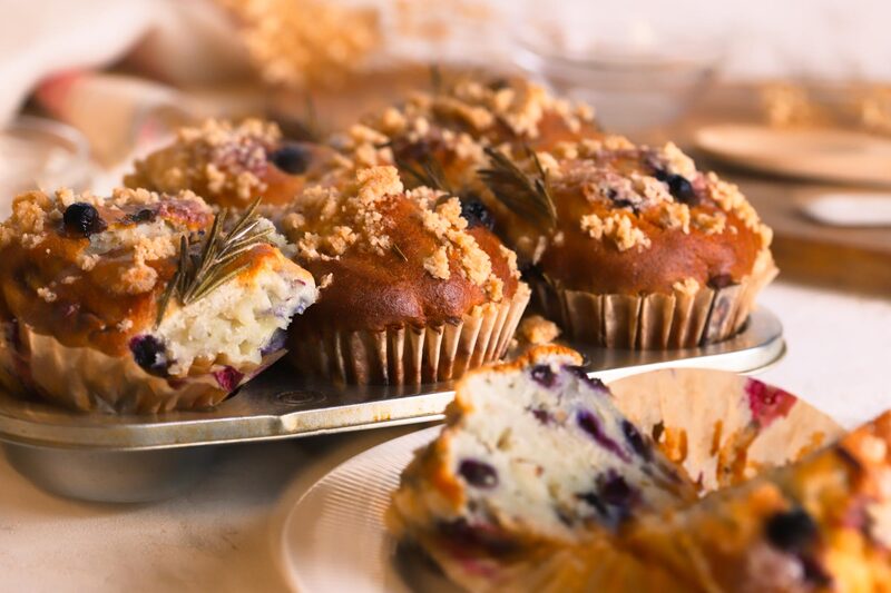 Gluten-free vegan blueberry rosemary muffins with streusel tops, one split open to show a moist crumb studded with blueberries, garnished with fresh rosemary