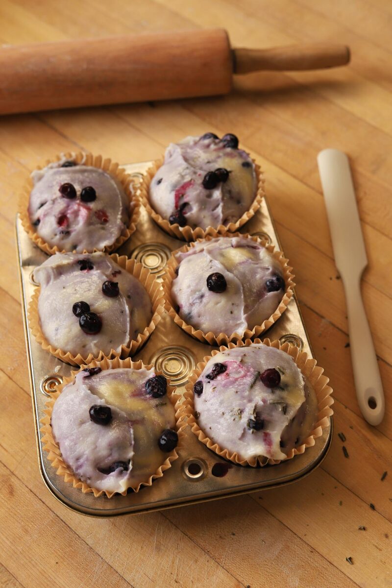 Unbaked blueberry muffin batter portioned into a lined muffin tin, dotted with blueberries and flecks of rosemary