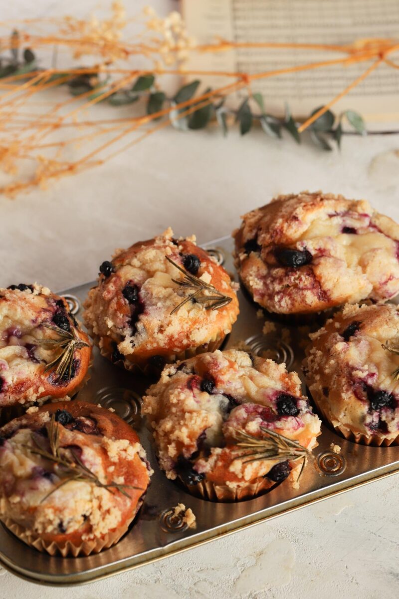 Six blueberry rosemary streusel muffins in a metal muffin tin, each topped with a sprig of fresh rosemary, on a styled table with dried flowers and eucalyptus