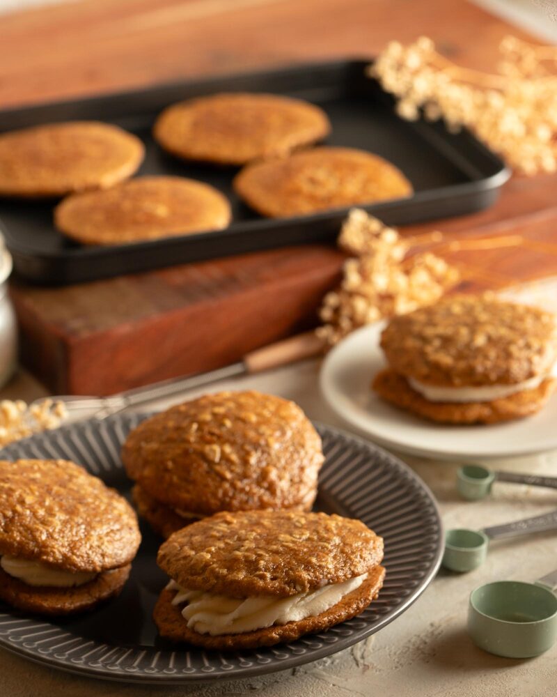 Golden-brown oatmeal cream pies stacked on a plate with measuring spoons nearby