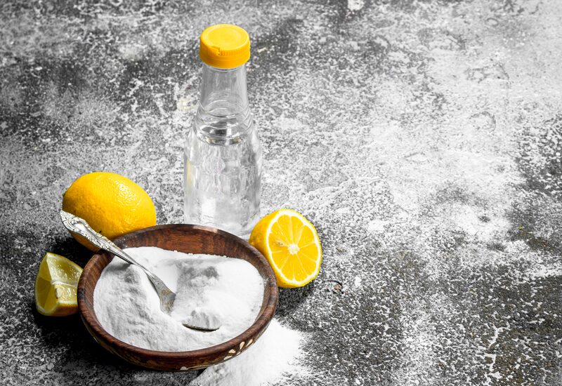 A bowl of baking soda with fresh lemons and a bottle of vinegar on a grey surface