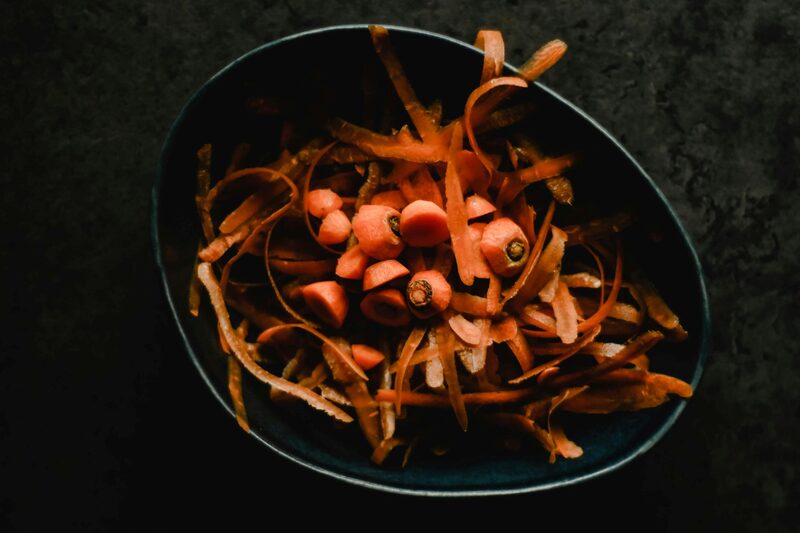 A dark bowl of carrot peels and trimmed carrot tops on a slate background