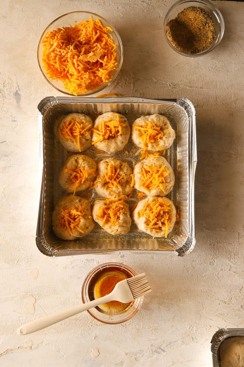 Proved dough balls in a pan, each dimple filled with shredded vegan cheese and sprinkled with garlic and herbs