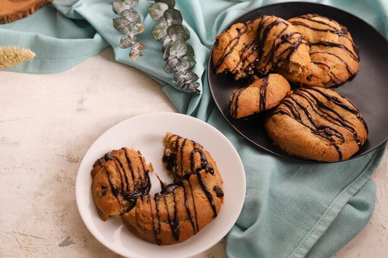 A chocolate-drizzled lava cookie pulled open on a white plate beside a black plate of whole cookies