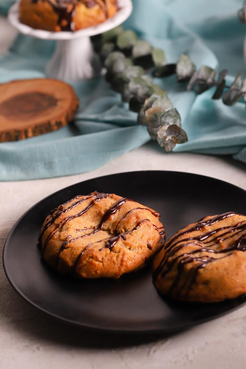 Two thick chocolate chip lava cookies with chocolate drizzle on a black plate