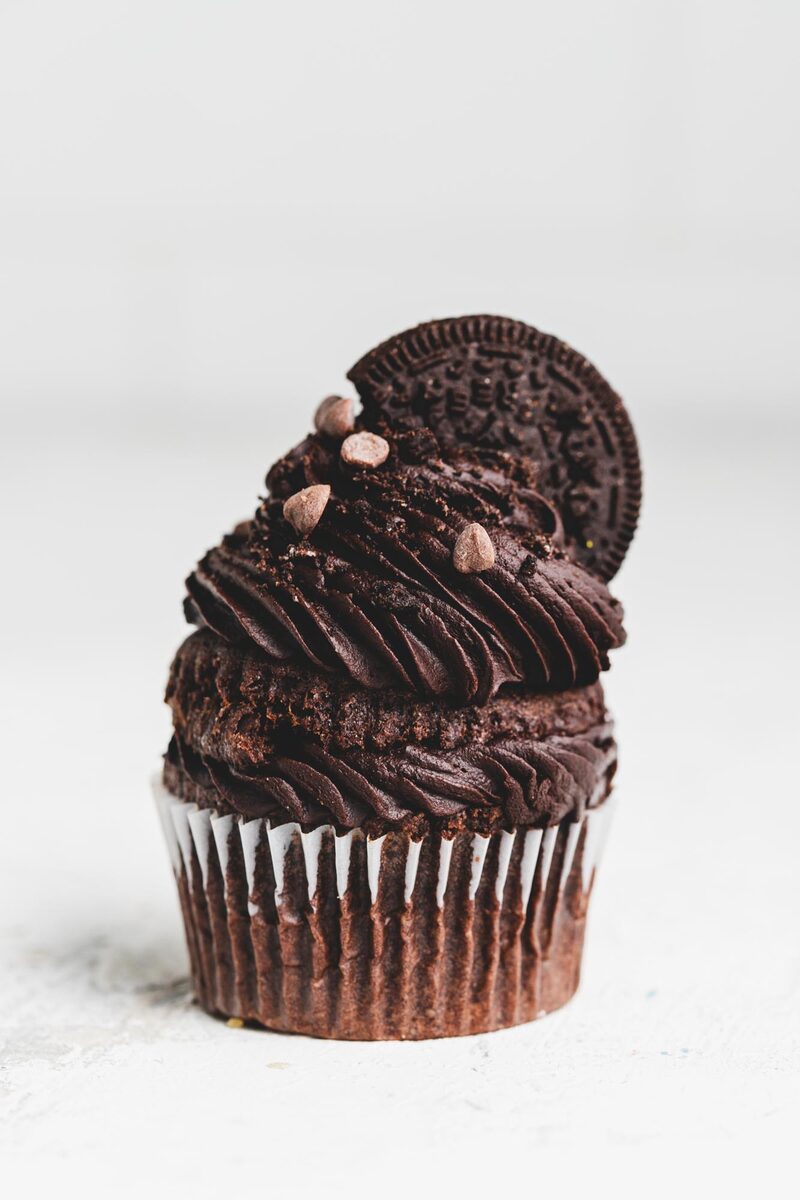 A chocolate cupcake piped with a tall swirl of chocolate frosting, topped with a cookie and cacao nibs