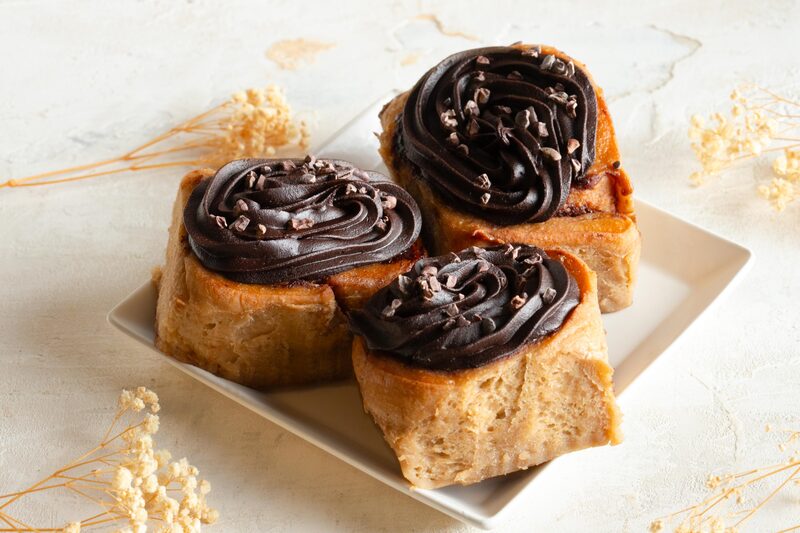 Three cinnamon rolls topped with swirls of dark vegan chocolate frosting and cacao nibs