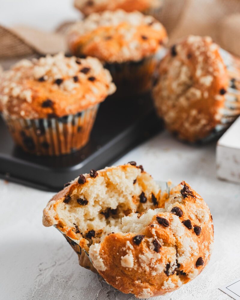 A chocolate chip muffin torn open to show a fluffy, tender interior, with more muffins behind it