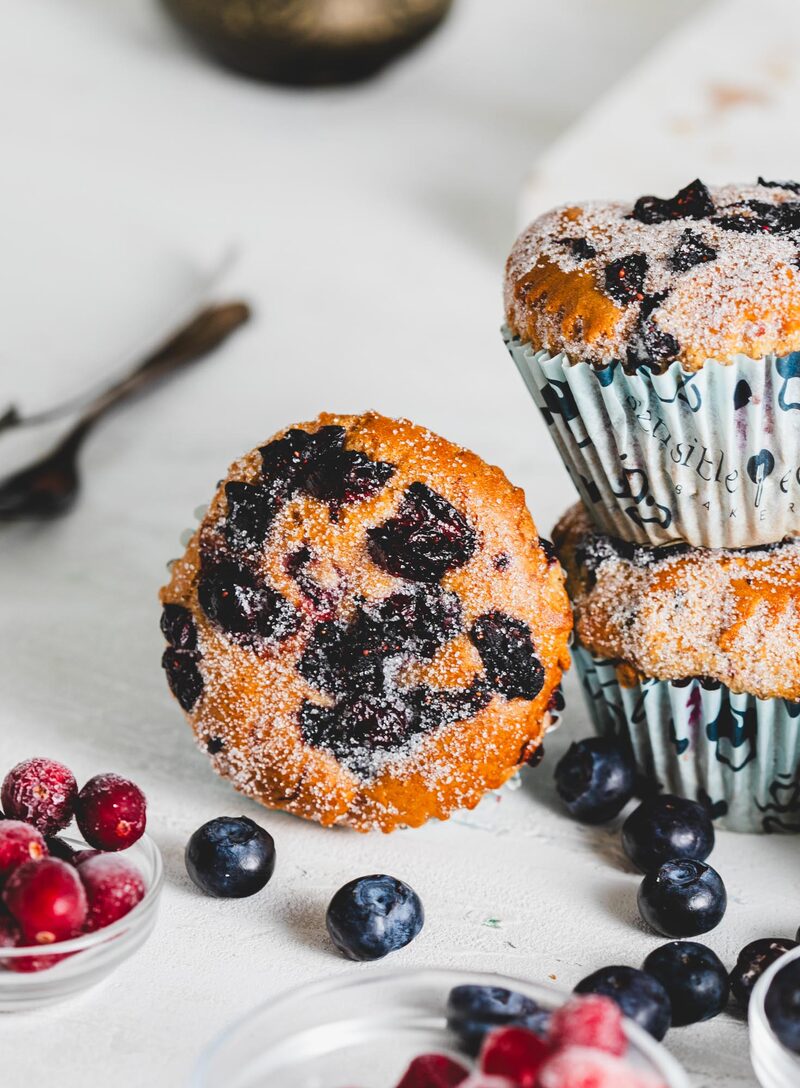 Sugar-topped blueberry muffins in Sensible Edibles wrappers surrounded by fresh blueberries and cranberries