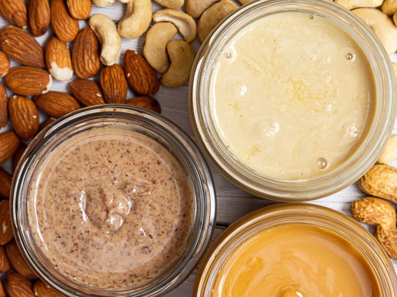 Open jars of almond, cashew, and peanut butter surrounded by scattered almonds, cashews, and peanuts