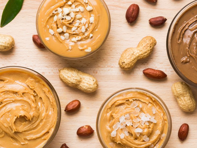 Small glass bowls of different nut butters on a wood board, topped with chopped nuts and flaky salt, with peanuts scattered around
