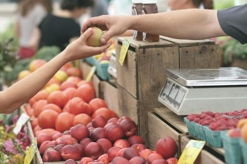 A shopper handing an apple to a farmers' market vendor over crates of nectarines and raspberries
