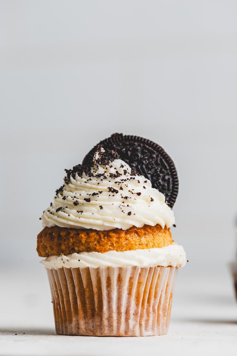A cupcake with swirled vanilla frosting, cookie crumbs, and a chocolate sandwich cookie on top