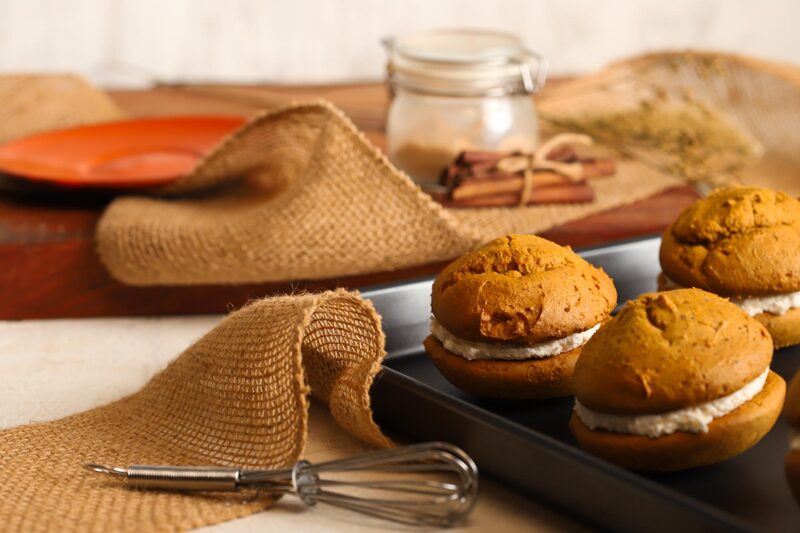 Pumpkin whoopie pies with cream filling on a baking tray, with burlap and a whisk alongside