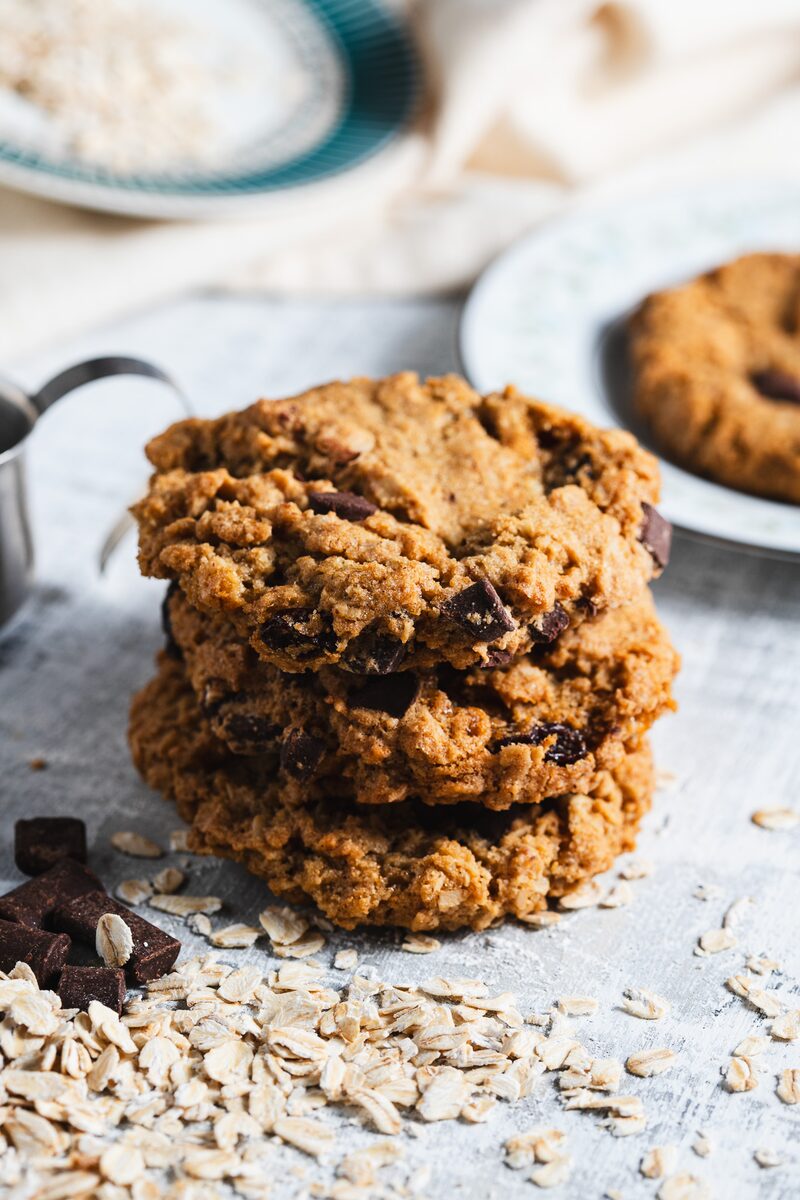 A stack of chocolate chip oat cookies with rolled oats and chocolate chunks scattered around