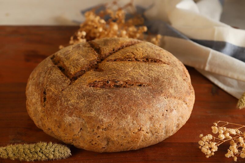 A rustic round loaf of gluten-free sourdough bread on a wooden board with dried grasses and a linen cloth