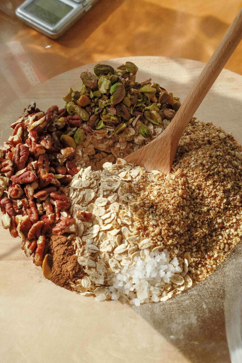 A wooden bowl of oats, pecans, pistachios, and seeds being stirred with a wooden spoon
