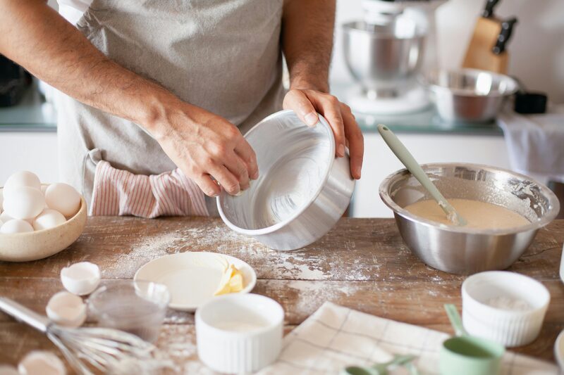 Baker dusting a buttered round cake pan with flour at a floured wooden worktable