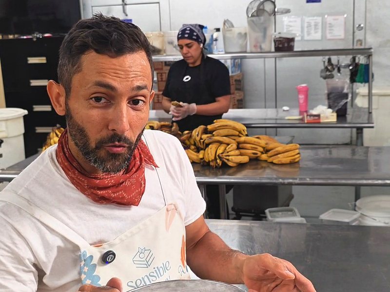 Wassem in a Sensible Bakery apron sifting baking soda through a fine-mesh sieve into a bowl