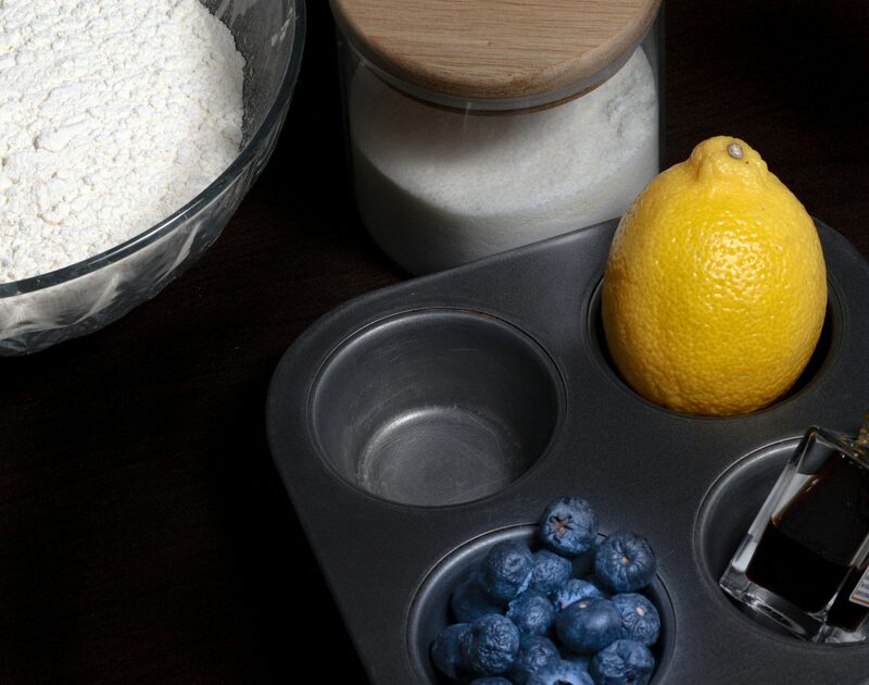 A lemon and blueberries sitting in a muffin tin next to bowls of flour and sugar