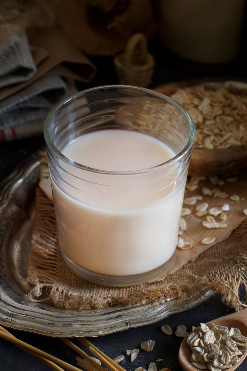 A glass jar of homemade oat milk beside a bowl of rolled oats and a jug pouring into coffee