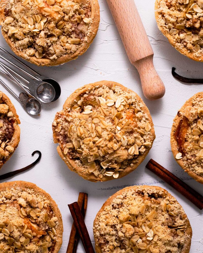 Mini apple crumble pies with oat-and-almond topping arranged with cinnamon sticks and vanilla pods