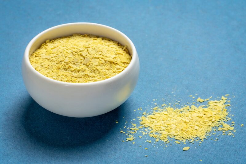 A small white bowl of yellow nutritional yeast flakes with some flakes spilled beside it on a blue background