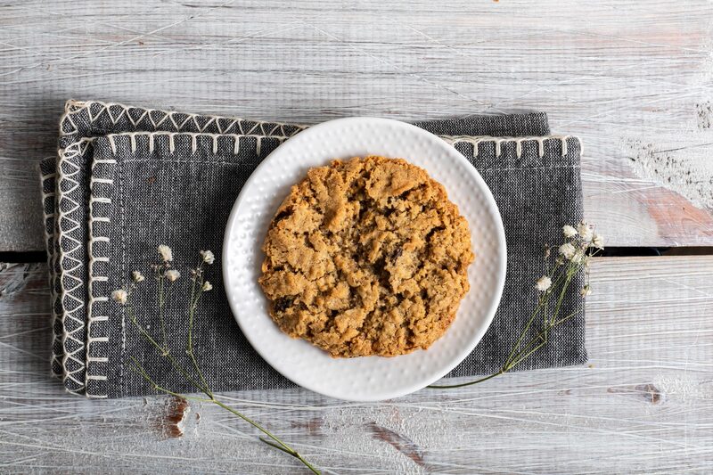 Overhead view of a single flattened oatmeal raisin cookie on a white plate set on a grey linen napkin