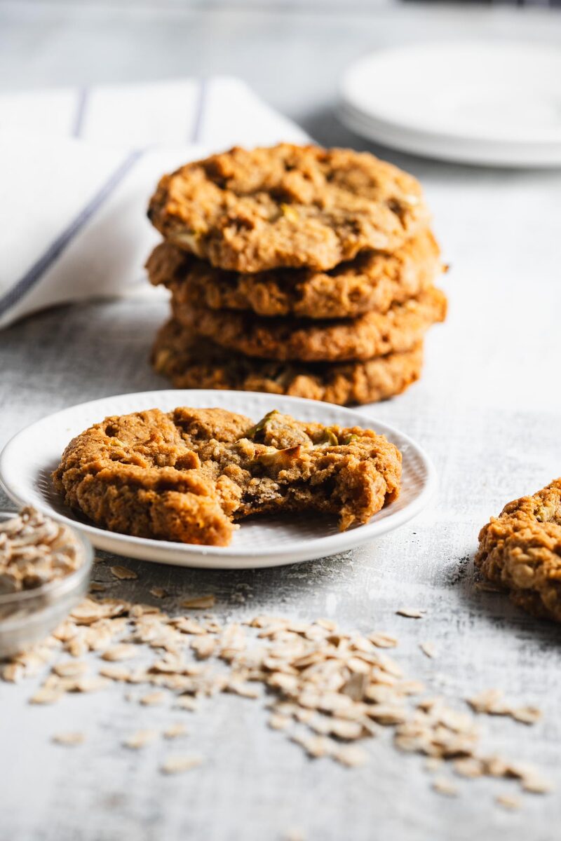 A stack of thick oatmeal raisin cookies behind a plated cookie with a bite taken out, loose oats scattered across the counter