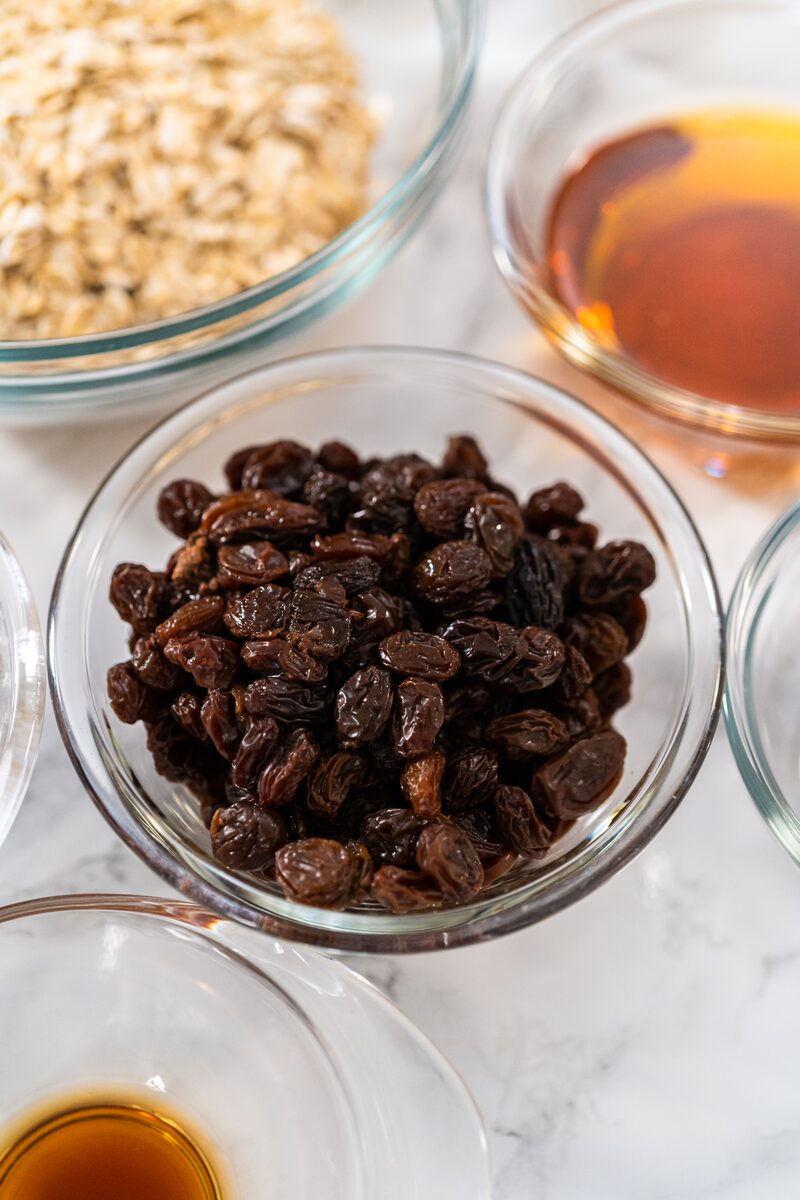 Glass bowls of rolled oats, raisins, maple syrup, and vanilla arranged on a marble counter