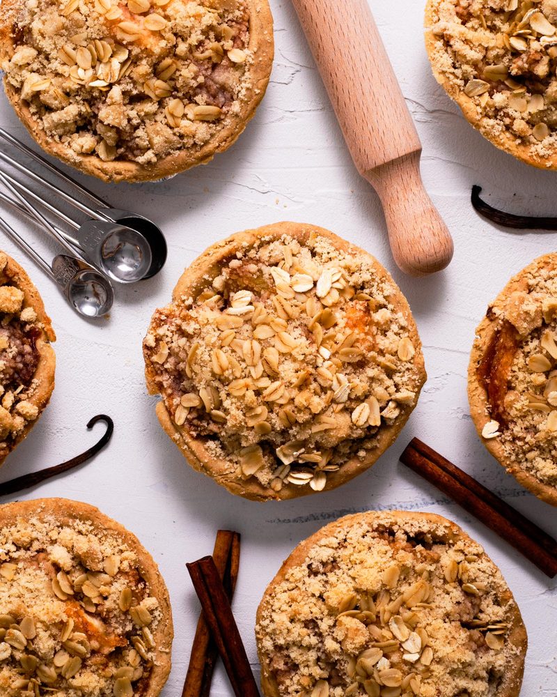 Several mini gluten-free vegan pies with golden oat-crumble tops, shown from above with a rolling pin, measuring spoons, cinnamon sticks, and vanilla beans
