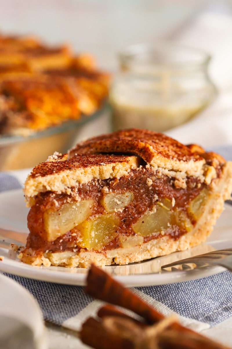 A thick slice of gluten-free vegan apple pie on a plate, the crust holding a clean cut, with a crumble top and cinnamon sticks alongside