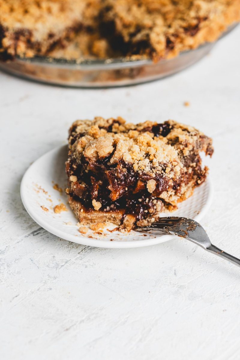 A plated slice of gluten-free vegan pie with a soft fruit filling and crumble top, fork resting alongside, the whole pie behind it