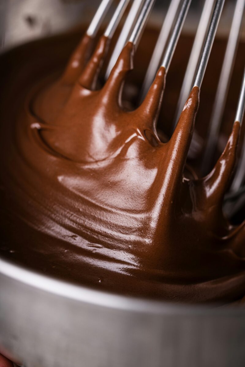 A whisk lifting glossy bloomed cocoa paste from a metal mixing bowl