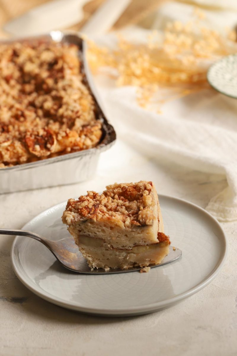 A square of gluten-free banana maple cake with a crumb top, plated on a fork with the pan behind, showing banana layered through the middle
