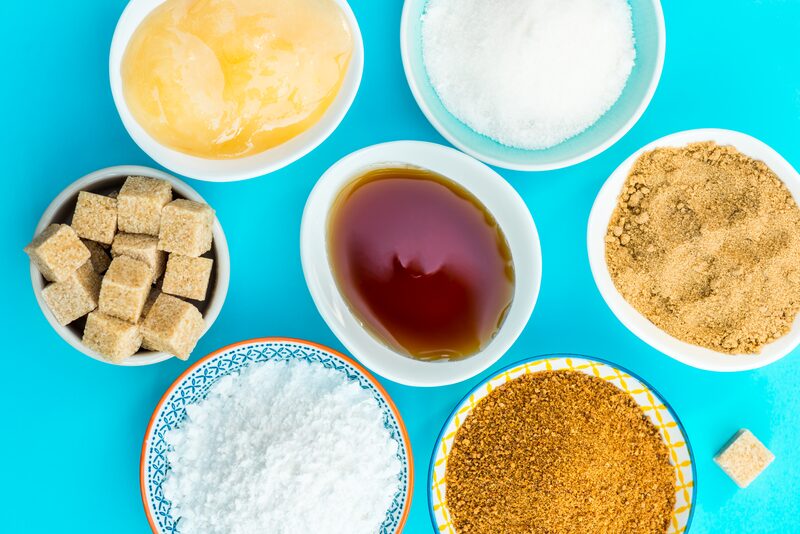 Bowls of honey, granulated sugar, maple syrup, brown sugar, sugar cubes, powdered sugar, and coconut sugar on a blue background