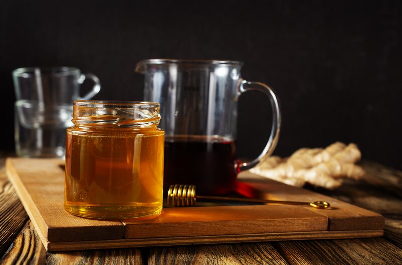 A jar of honey and a pitcher of dark syrup on a wooden board with fresh ginger