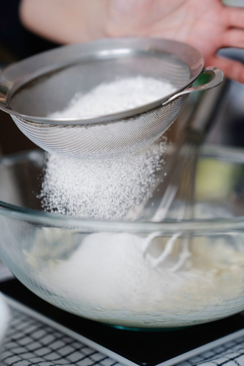 Flour being sifted through a fine mesh sieve into a glass mixing bowl