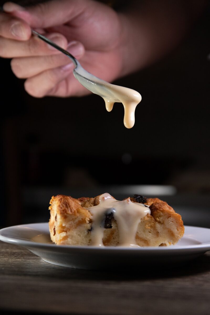 A spoon drizzling glossy sweetened condensed milk over a slice of bread pudding
