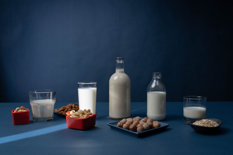 A lineup of plant-based milks in glasses and bottles beside almonds, cashews, hazelnuts, and oats