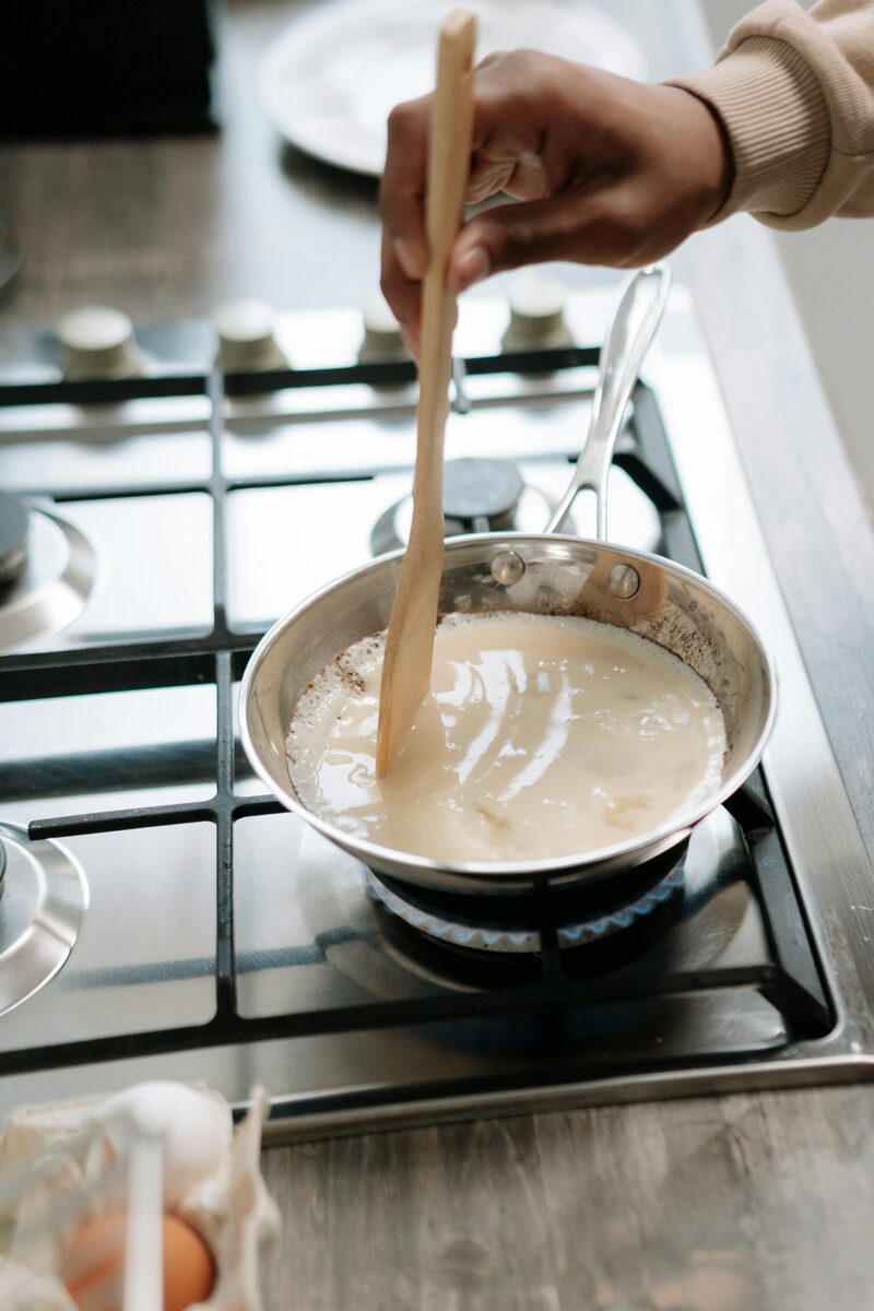 A creamy plant-milk-and-sugar mixture reducing in a saucepan, stirred with a wooden spatula
