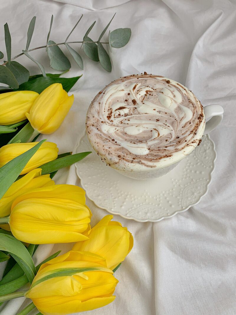 A latte topped with swirled vegan cream foam dusted with cocoa, beside yellow tulips